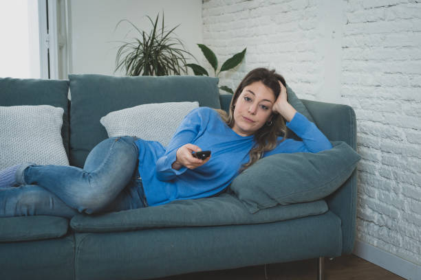 Bored woman watching TV helpless in self Isolation at home during mandatory lockdown due to coronavirus outbreak. Young upset woman on sofa using control remote zapping bored of TV and sedentary life. stock photo