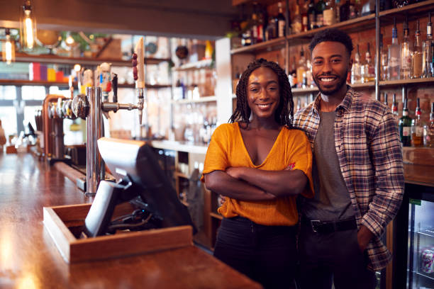 Portrait Of Smiling Couple Owning Bar Standing Behind Counter Portrait Of Smiling Couple Owning Bar Standing Behind Counter black people bar stock pictures, royalty-free photos & images