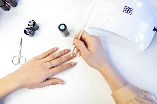 Young woman doing her own manicure at home. Using UV/LED nail lamp, scissors, nail files and different colors of nail polish. Drawing some details in different colors on nails.