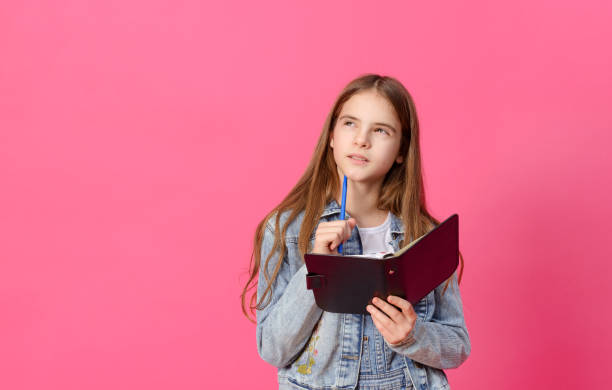 1 cute pensive white girl 10 years old in a blue denim jacket with a notebook and pen on a pink background - 10 11 years child human face female imagens e fotografias de stock