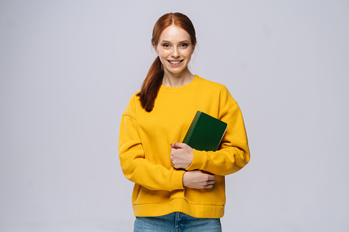Portrait of cheerful young woman college student holding book and looking at camera on isolated gray background. Pretty redhead lady model wearing casual clothes emotionally showing facial expressions