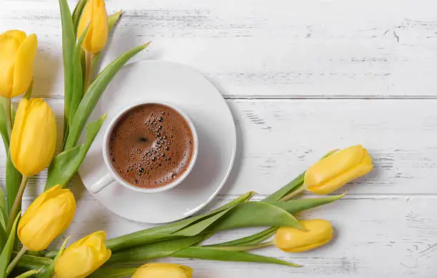 Photo of lots of yellow tulips, a cup of black coffee on a white wooden background, gift, flowers, top view
