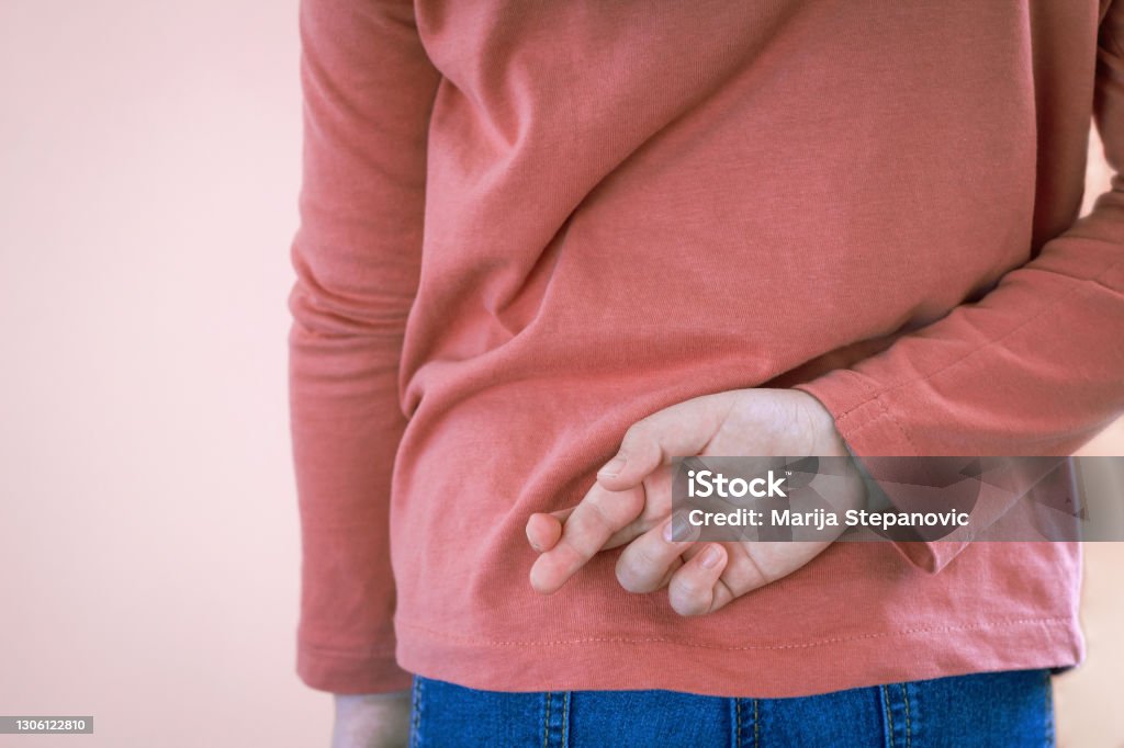 Lying concept with girl child crossing her finger behind back on light background with copy space. April Fool's Day. Female child  crossing her fingers behind  the back on light background with copy space. Lie and cheating, lying concept. April Fools' Day Dishonesty Stock Photo