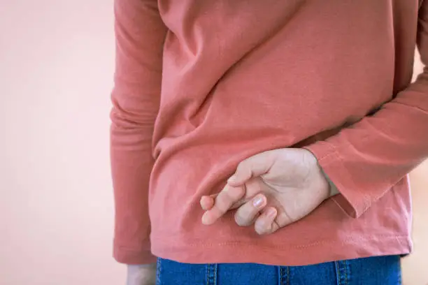 Photo of Lying concept with girl child crossing her finger behind back on light background with copy space. April Fool's Day.