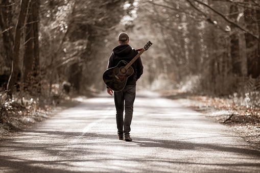 Color image depicting the rear view of a man walking on a beautiful tree lined country road. He is enjoying the freedom of exploring and wandering in nature, and he has an acoustic guitar strapped to his back. Room for copy space.