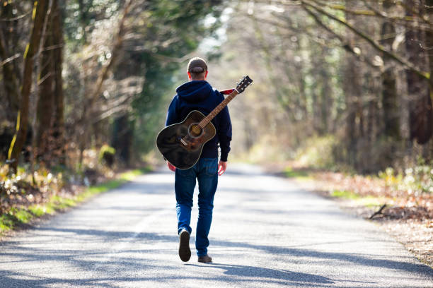 istock Man walking on country road with guitar on his back 1306124108