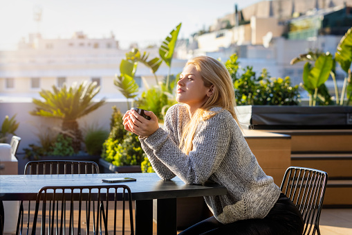 A young, blonde woman enjoys her morning coffee outdoors on the terrace under warm sunlight