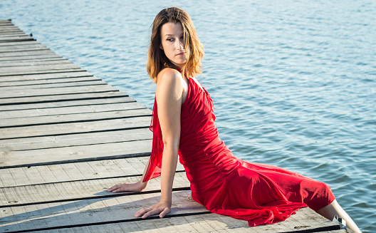 Young brunette Caucasian woman wearing elegant red dress is sitting on a wooden jetty on a lake on a sunny summer afternoon.