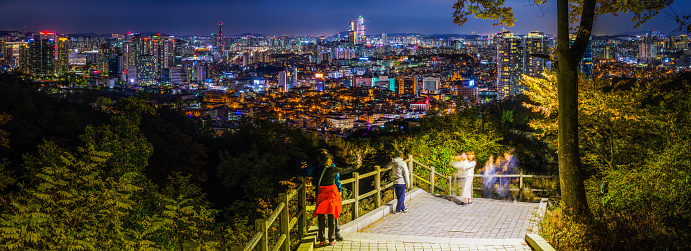 Tourists taking selfies on the steps up Namsan Mountain overlooking the neon night skyscraper cityscape of central Seoul, South Korea’s vibrant capital city.