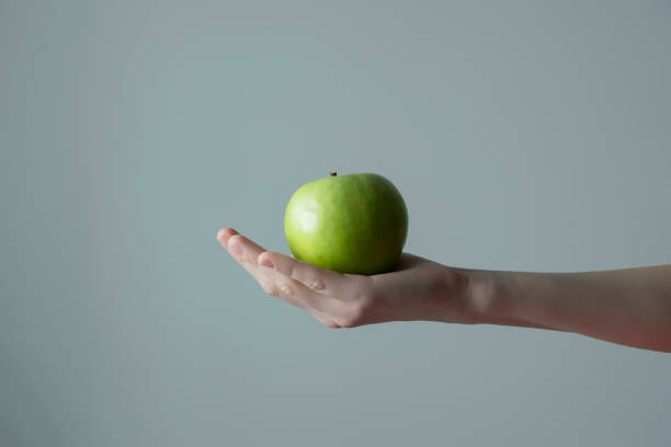 istock Woman`s hand holding green apple grey neutral background - raw photo. 1306154384