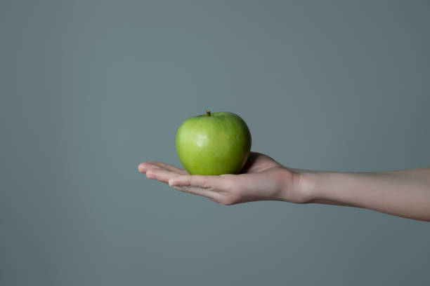istock Woman`s hand holding green apple grey neutral background - raw photo. 1306155381