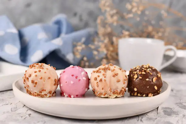 Small colorful cake balls glazed with white, pink and brown chocolate with sprinkles in a row on white plate