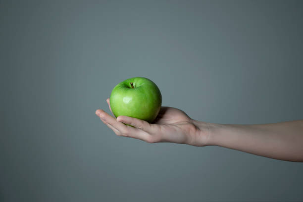 istock Woman`s hand holding green apple grey neutral background - raw photo. 1306155628
