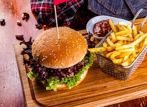Hamburger with basket of French fries on and tomato sauce on wooden cutting board