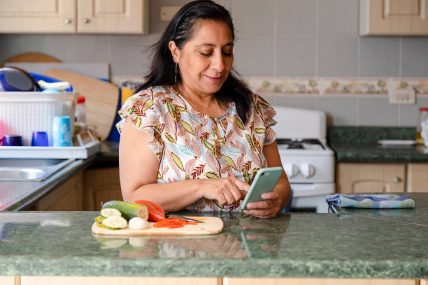 maman hispanique recherchant des recettes sur sa femme téléphone-mûr préparant la salade saine et organique tout en vérifiant sa cuisine de téléphone portable-femme au foyer tandis qu’elle regarde un téléphone - stereotypical homemaker photos et images de collection