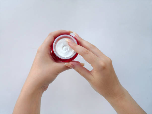 pote aberto vermelho com produto cosmético branco (rosto ou creme corporal) nas mãos de uma jovem com pele perfeita e unhas longas (baby boomer ou coloração de fade francês) em fundo branco - fade in - fotografias e filmes do acervo