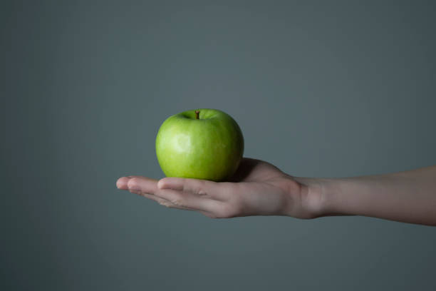 istock Woman`s hand holding green apple grey neutral background - raw photo. 1306192125