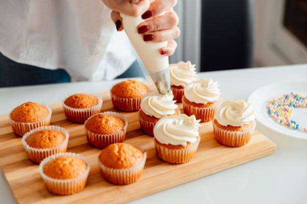 frau schmückt frisch gebackene cupcakes mit sahne - zuckerguß stock-fotos und bilder