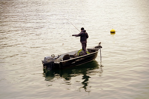 Zurich, Switzerland, - 02 06 2021: A hobby fisherman fishing out the hook with a fish out of lake Zurich in winter. He is standing in a boat and there is a yellow buoy on the water surface around him.