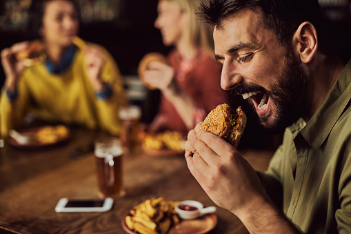 Hungry man biting hamburger while having lunch with friends in a pub.