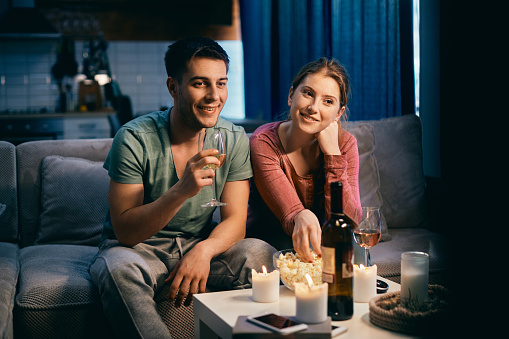 Smiling couple drinking wine and eating popcorn while watching movie on TV at night at home.