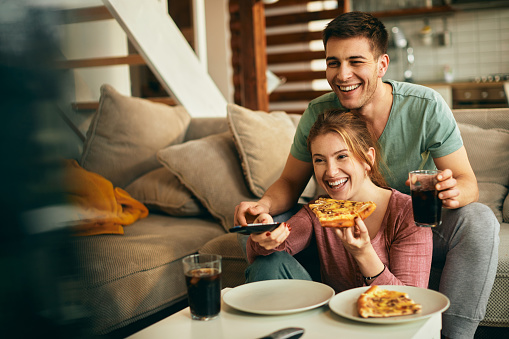 Young couple having fun while eating pizza and watching TV in the living room.