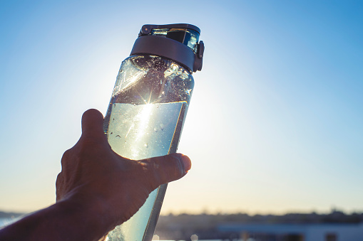 Man holding up a grey reusable water bottle in the sunshine. The bottle is full with a blue sky background