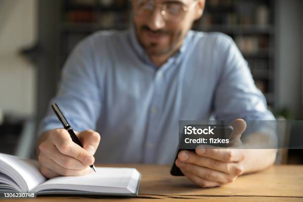 Geschäftsmann Sitzt Am Schreibtisch Halten Smartphone Schreibt Zeitplan In Veranstalter Stockfoto und mehr Bilder von Kalender