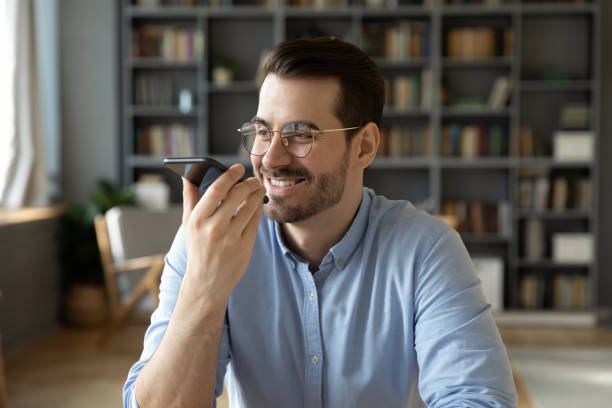 笑顔のビジネスマンは、スマートフォンを持ってオフィスに座って声のメッセージを残します - 会議用電話 ストックフォトと画像