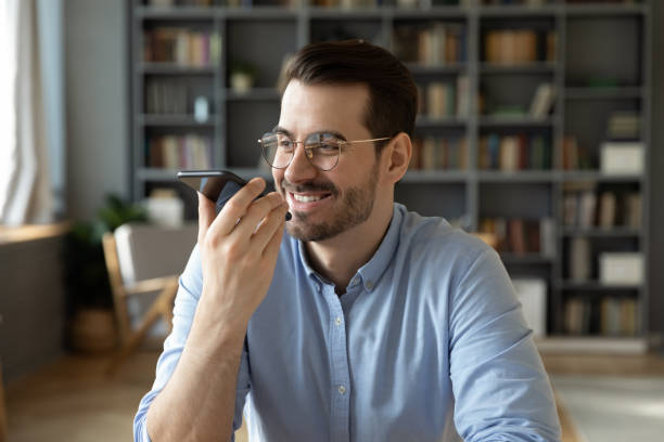 uśmiechnięty biznesmen siedzi w biurze trzymając smartfon pozostawia wiadomość głosową - to do list audio zdjęcia i obrazy z banku zdjęć