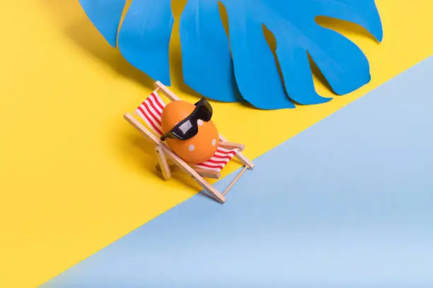 Photo of Creative funny composition with Easter egg with sunglasses while sitting on deck chair on illuminating yellow and pastel blue background