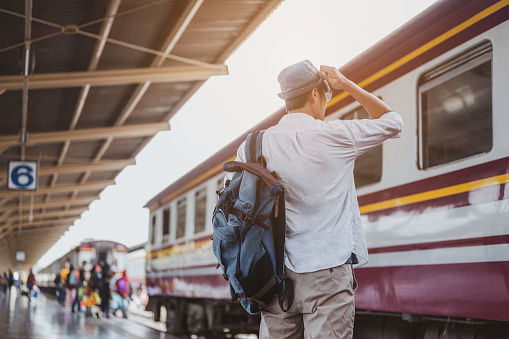 Asian man traveler with backpack waiting for train, Asian backpacker with hat standing on railway platform at Bangkok train station. Holiday, journey, trip and summer Travel concept.