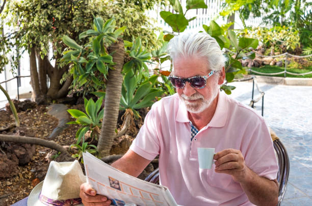 istock Attractive senior man with sunglasses sitting at the coffee table while reading the newspaper and drinking a coffee cup - around him a tropical garden 1306256632