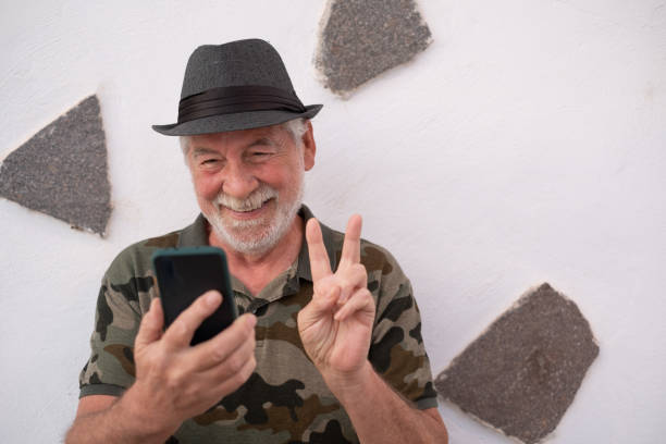 istock Smiling senior man with hat using smart phone standing outdoors close to a white wall, in video call. Older people enjoying new technology 1306256952