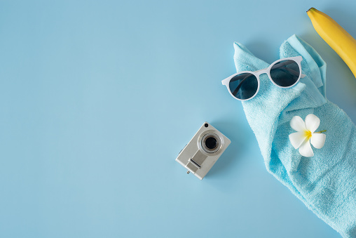 lat put travel accessories in the new normal style with cameras, glasses, bananas and a towel placed on a blue background.