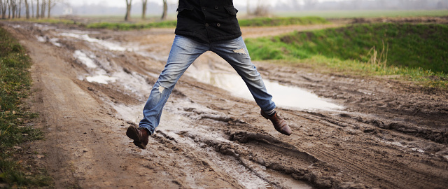 A man in shoes jumps over a muddy puddle outside the city.