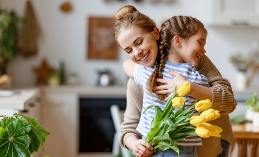 Loving little daughter hugging happy mom and greeting with bouquet of yellow tulips during Mothers day celebration at home