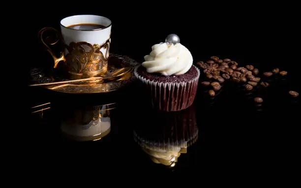 Cupcake on black glass background with reflection, golden coffee cup. Homemade cake with cream and roasted coffee beans. Selective focus.
