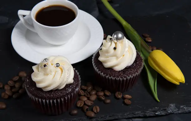 Two cupcakes on black background and white coffee cup, homemade cake with cream and roasted coffee beans.Selective focus, blur background.