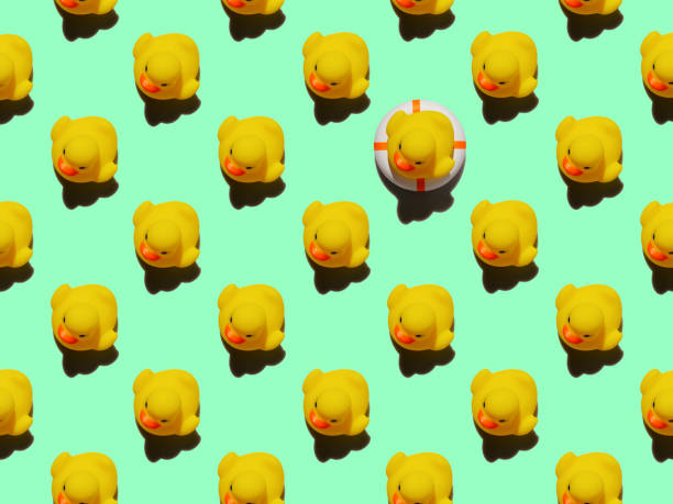istock Many yellow rubber ducks in an abstract formal repeating pattern with one yellow rubber duck sitting on an inflatable life belt standing out from the crowd, set on a turquoise background. 1306260773