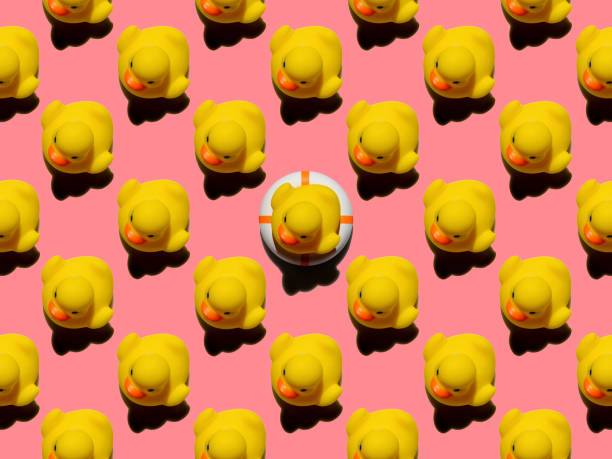 istock Many yellow rubber ducks in an abstract formal repeating pattern with one yellow rubber duck sitting on an inflatable life belt standing out from the crowd, set on a pink background. 1306260786