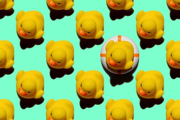 istock Many yellow rubber ducks in an abstract formal repeating pattern with one yellow rubber duck sitting on an inflatable life belt standing out from the crowd, set on a turquoise background. 1306260787