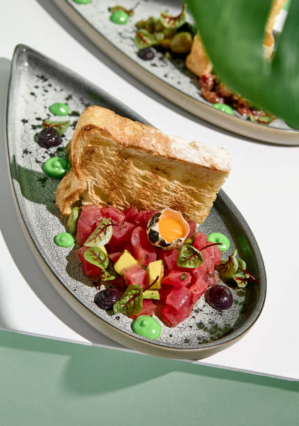 Tuna tartar with quail egg and toast bread. Raw fish tartare restaurant appetizer on white table with green wall. Day sunlight with hard shadow of monstera palm leaves. Summer or spring food concept tartar de atún con huevo de codorniz y pan tostado. aperitivo de restaurante tartar de pescado crudo sobre mesa blanca con pared verde. luz del sol del día con sombra dura de hojas de palma monstera. concepto de comida de verano o primaveraÑ
