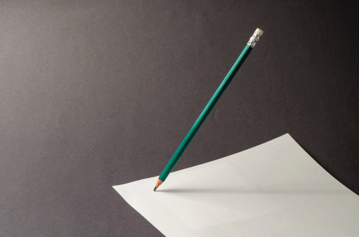Standing pencil and a sheet of paper on a gray background. A green pencil stands on a white blank sheet of paper. Selective focus. No people