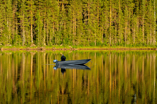 Ljusdal, Sweden, 6/26/2020.
Beautiful summer view of a small boat anchored in a river, with a man fishing. A pine forest in bright sunlight reflecting like a mirror in the water
