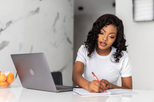 istock Smiling young woman entrepreneur sitting at a table in her kitchen writing down notes and working on a laptop 1306273732