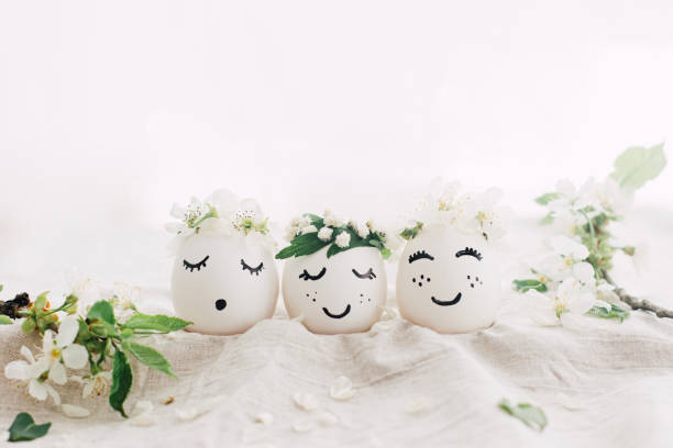 istock Natural easter eggs with drawn cute faces in floral wreaths on linen fabric with bloom in light 1306275686