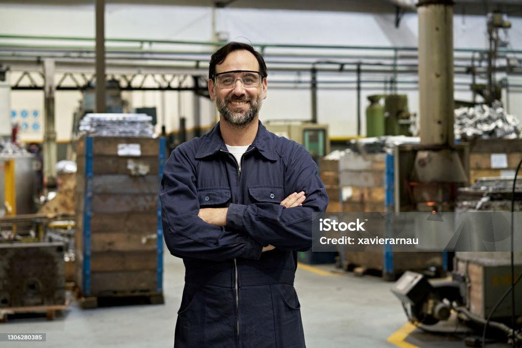 Portrait of Bearded Foundry Worker in Early 40s Partial front view of Caucasian man in coveralls wearing protective eyewear and standing on foundry floor with arms crossed smiling at camera. Coveralls Stock Photo