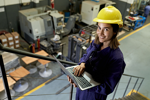 Elevated view of Caucasian woman in coveralls and work helmet standing on indoor staircase and pausing from typing to smile at camera.