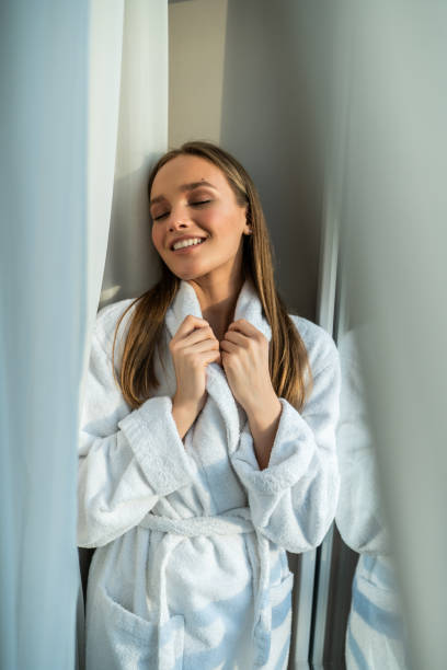 istock Attractive young woman in bathrobe looking at camera and smiling while sitting on a window sill in the bathroom 1306285857